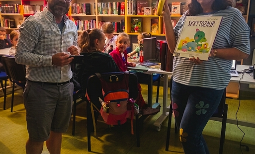 Grupa uśmiechniętych dzieci podczas zajęć z okazji Dnia Dziecka w Filii nr 4. Dzieci siedzą przy stolikach, układając puzzle z dinozaurami i trzymając w rękach kolorowe baloniki oraz pamiątkowe dyplomy. W tle widać biblioteczne regały z książkami ozdobion